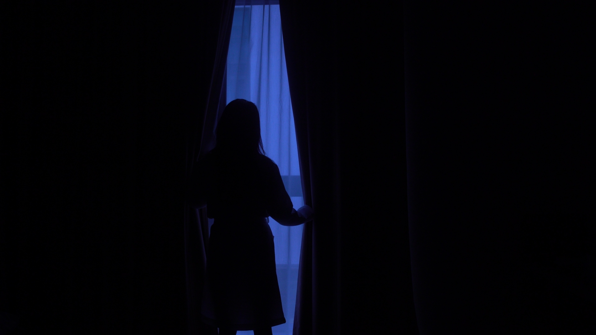A woman in silhouette parts the curtains of a dark hotel room.